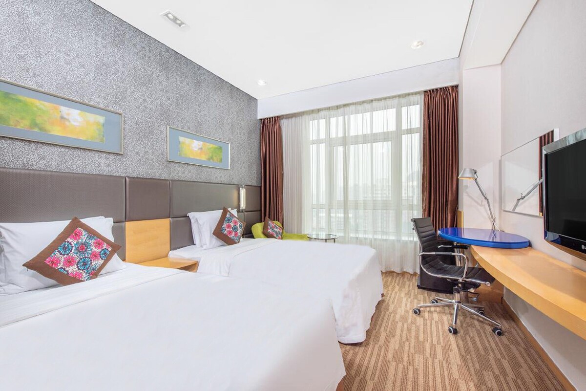 Quarto de hotel com duas camas, uma escrivaninha com computador, televisão e uma janela grande com cortinas.