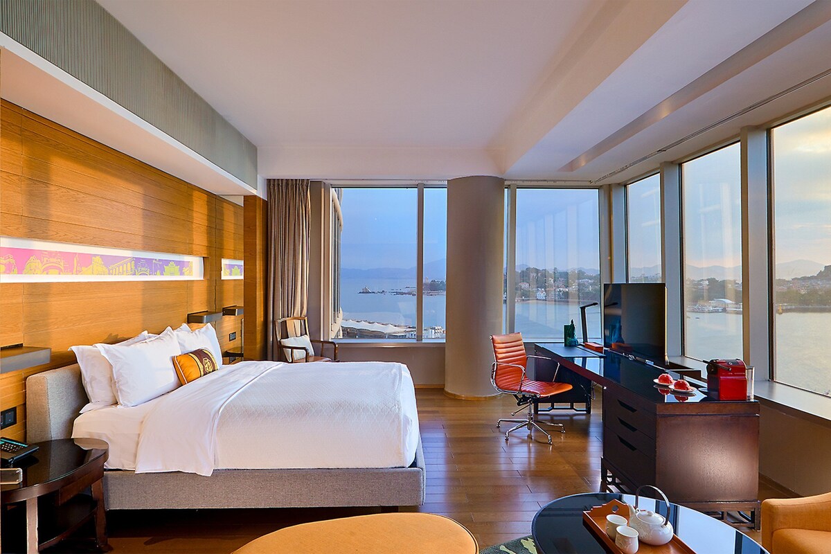 Een hotelkamer met een bed, bureau en grote ramen met uitzicht op een waterpartij en stadsgezicht.