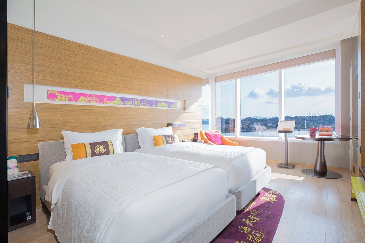 Een moderne hotelkamer met twee bedden, een groot raam met uitzicht op de stad, een kleine tafel en een decoratief wandobject.
