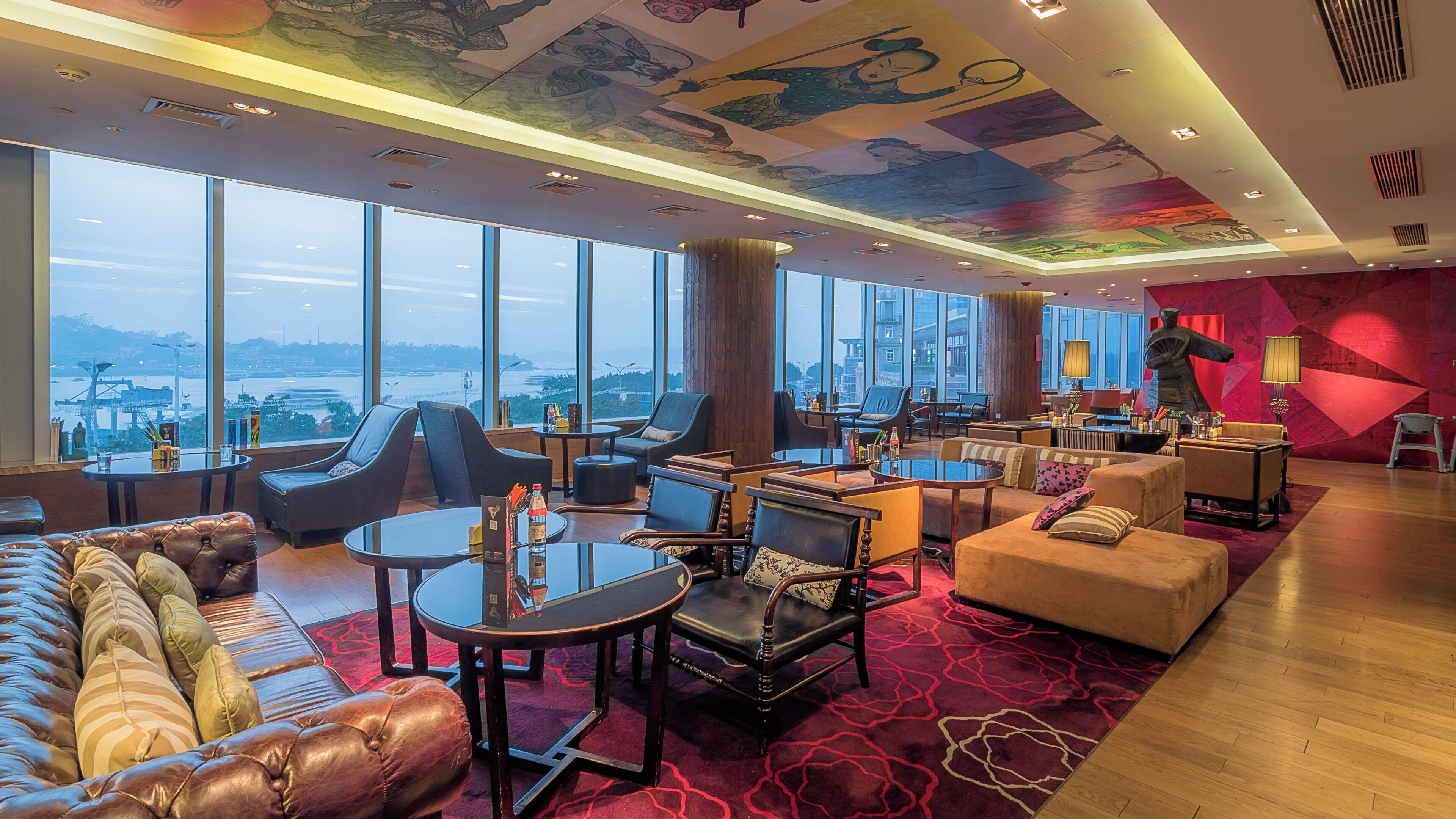 A spacious lounge area with leather sofas, armchairs, and tables, large windows offering a city view, and a vibrant ceiling mural.