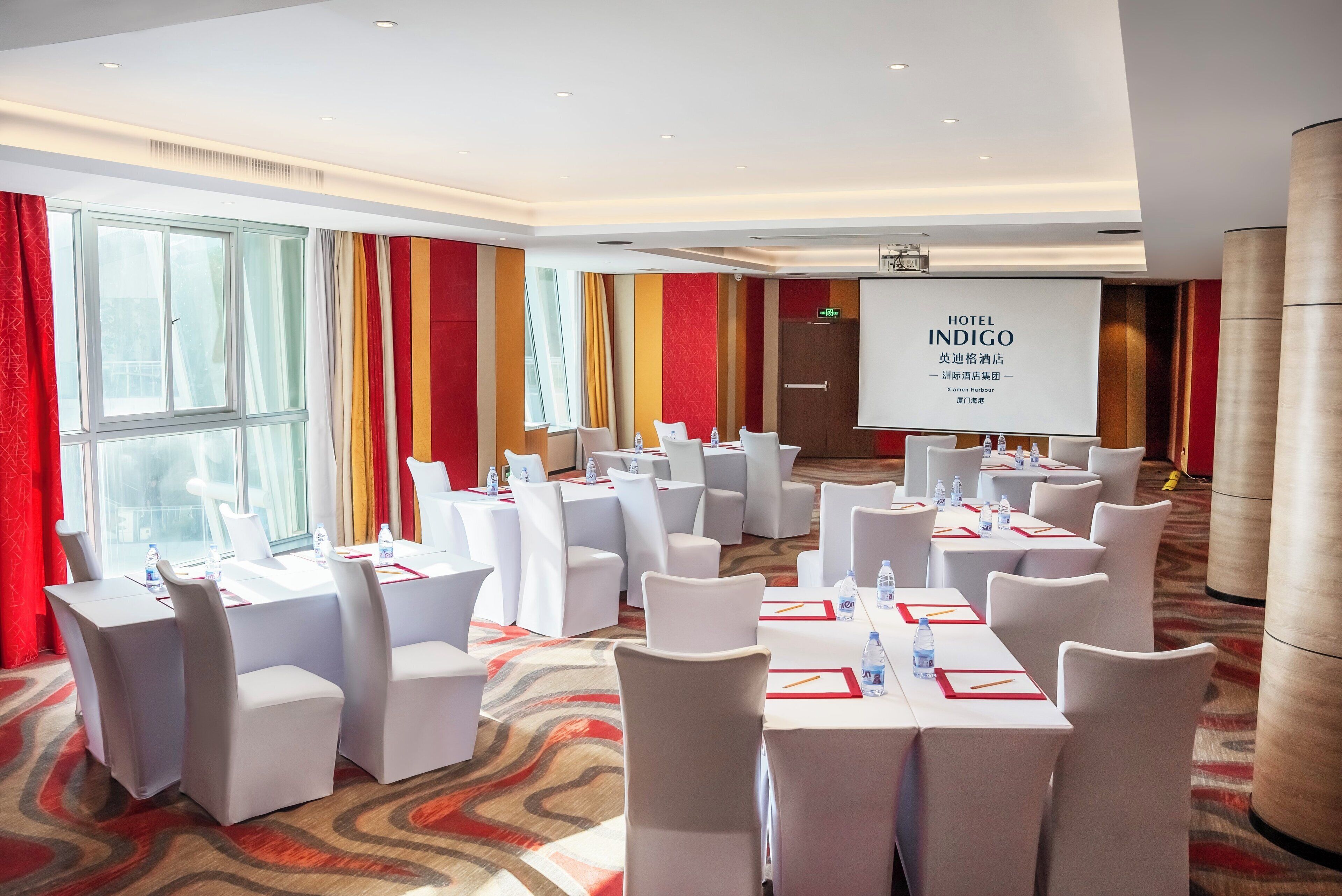 Un salón de conferencias con varias mesas y sillas, una pantalla de proyector y un amplio ventanal con cortinas.