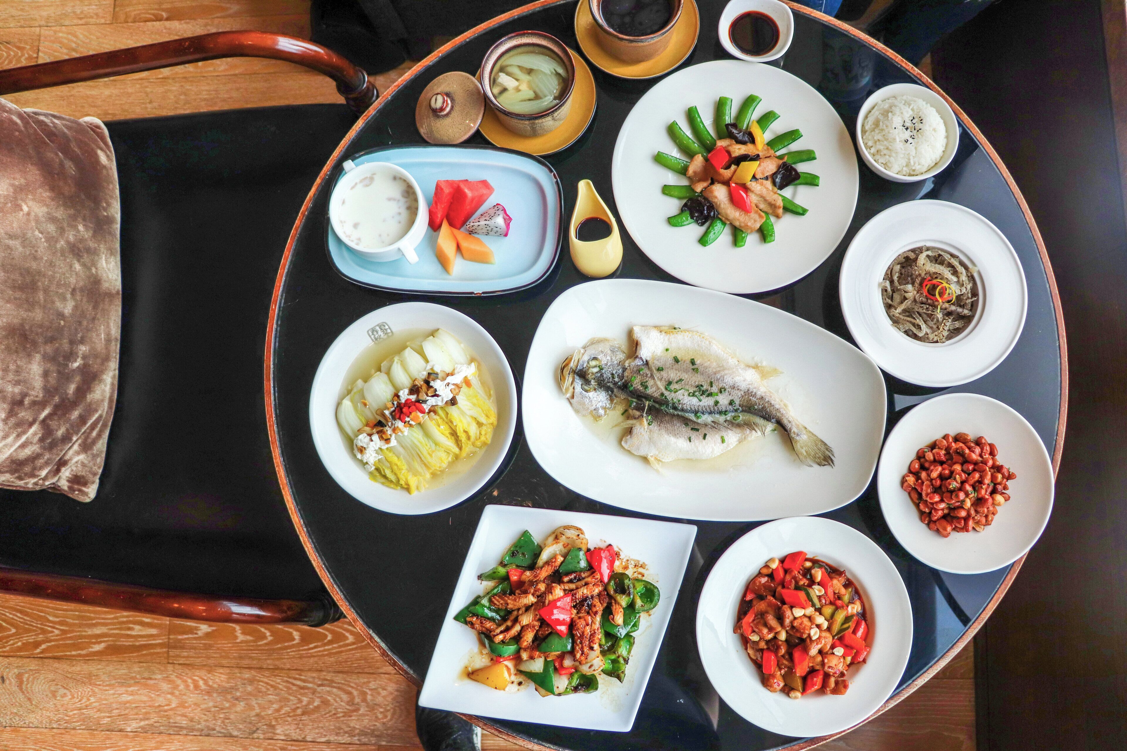 Une table ronde, dressée pour le dîner, avec différents plats, notamment du poisson, des légumes et du riz, ce qui laisse supposer qu’un repas est sur le point d’être servi.