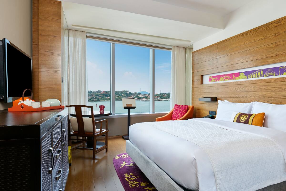Habitación de hotel con una cama grande, cabecera de madera, televisor, escritorio con silla y vista a un cuerpo de agua.