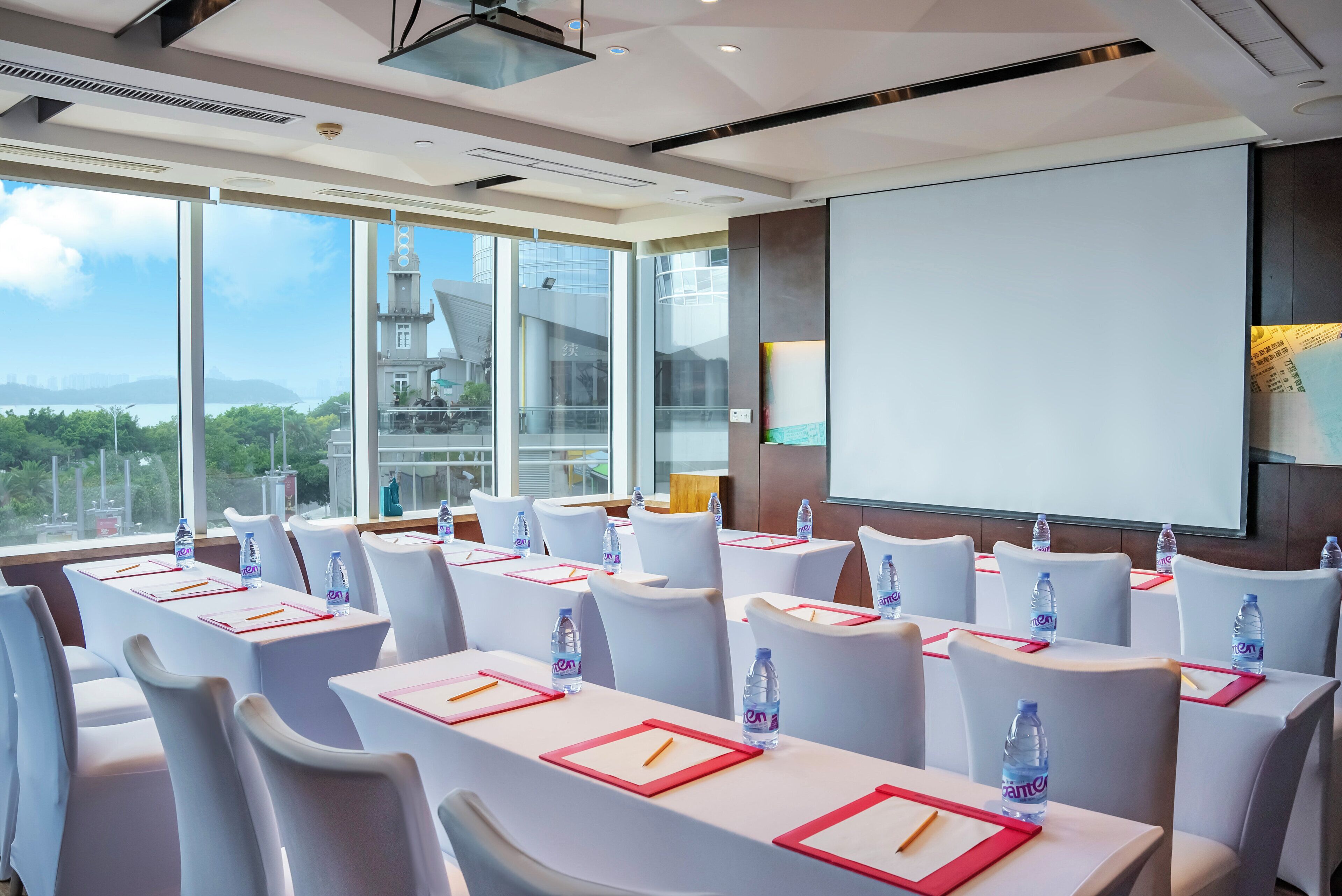 A conference room with tables set for meetings, equipped with water bottles and nameplates, large windows offering a cityscape view, and a projection screen.