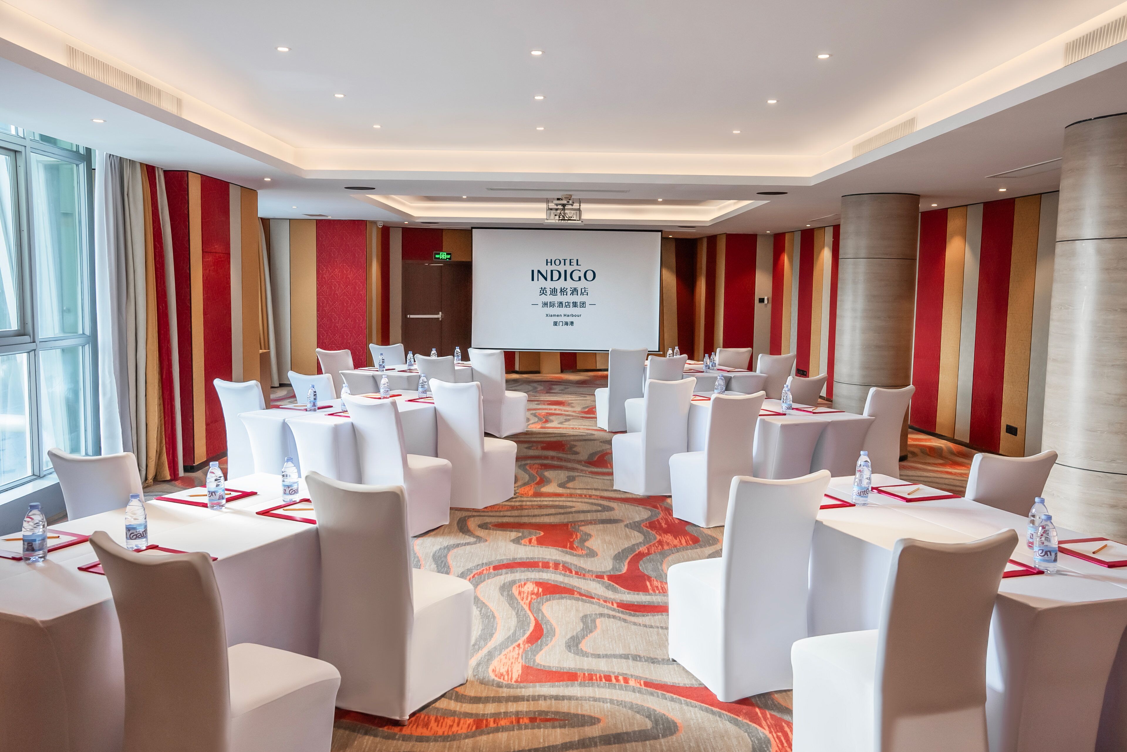 A conference room with white tables and chairs, a large screen displaying 'HOTEL INDIGO', and a patterned carpet.