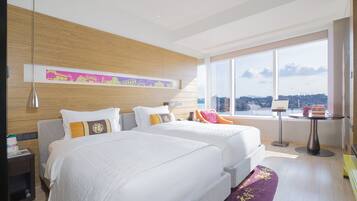 Habitación de hotel con dos camas, un ventanal grande con vista a la ciudad, una mesita y una mesita de noche con lámpara.