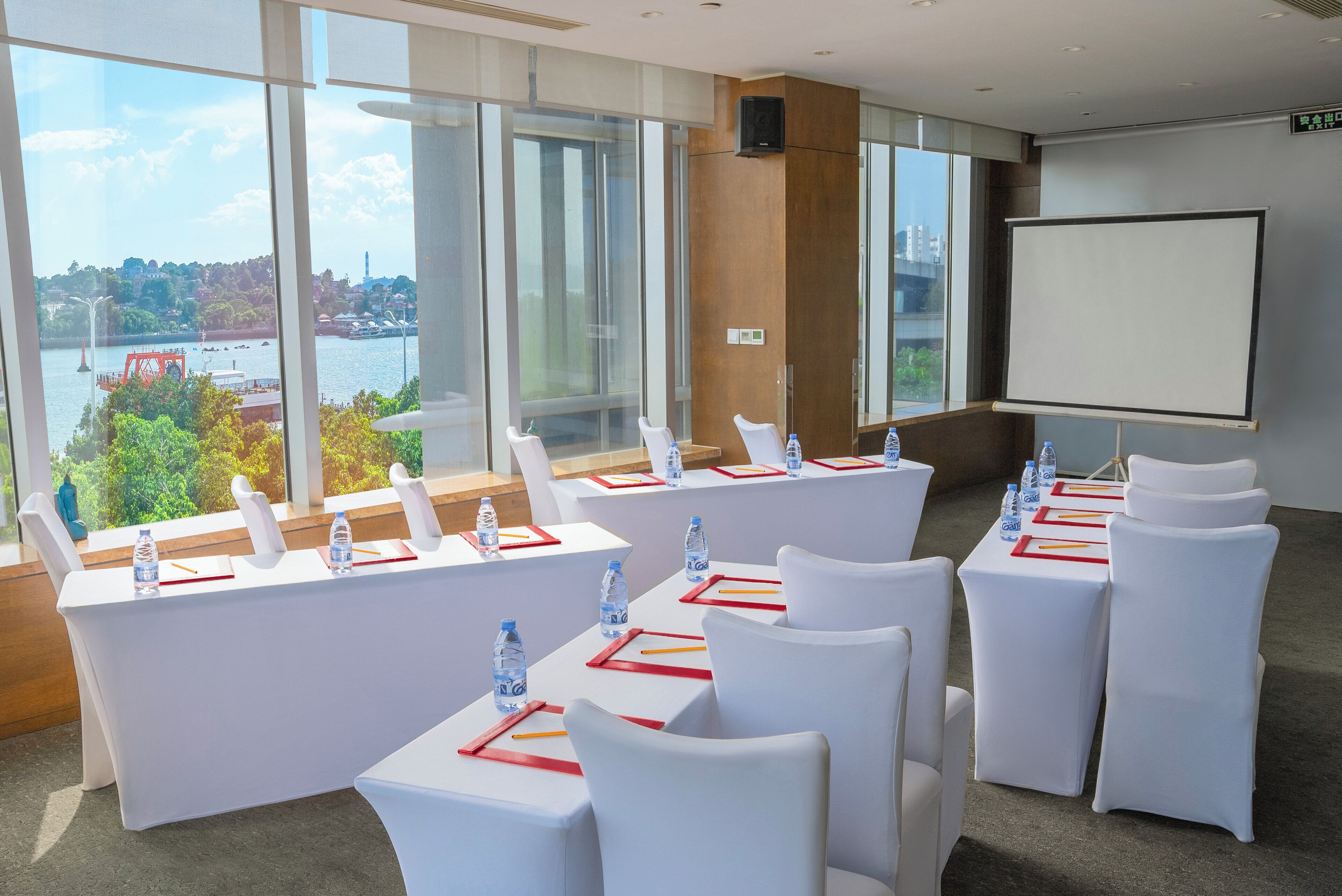 Ein Konferenzraum mit weißen Tischen und Stühlen, einer Projektionsleinwand und Blick auf einen Fluss sowie die Stadtlandschaft.