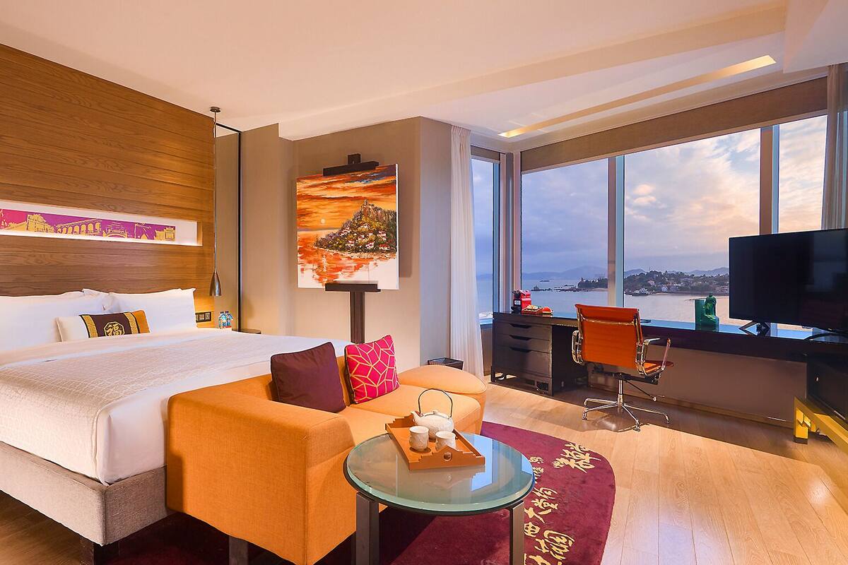 Habitación de hotel con cama, sofá, escritorio, televisor y un ventanal con vistas.
