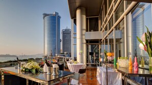 Una terraza en la azotea con vistas a modernos rascacielos, una mesa dispuesta con un jarrón de flores y diferentes botellas.