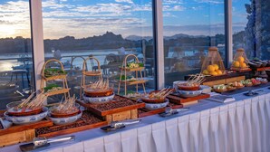 Taula de buffet amb diversos plats i utensilis, amb un paisatge idíl·lic al fons a través de grans finestres.