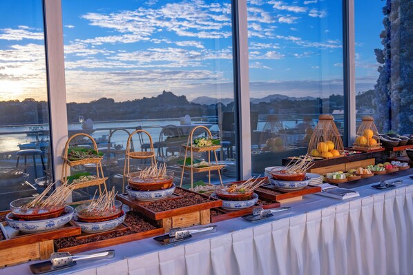 A buffet table with various dishes and utensils, set against a backdrop of a scenic view through large windows.