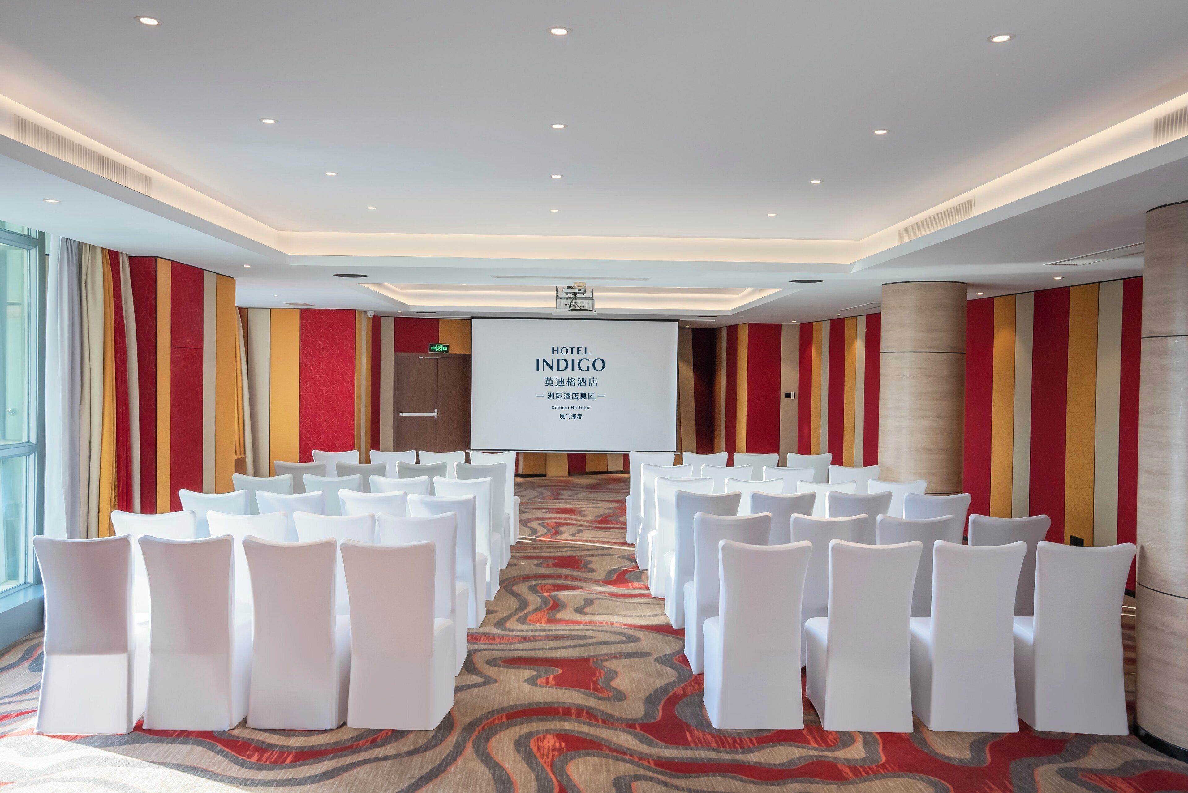 Un salón de conferencias con filas de sillas blancas, una pantalla grande que muestra 'HOTEL INDIGO' y suelo alfombrado.