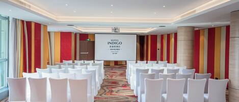 Un salón de conferencias con filas de sillas blancas, una pantalla grande que muestra 'HOTEL INDIGO' y suelo alfombrado.