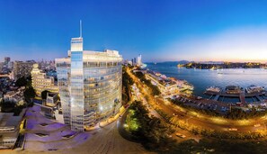 Un hôtel moderne à la façade en verre, situé près d’une promenade en bord de mer où sont amarrés des bateaux.