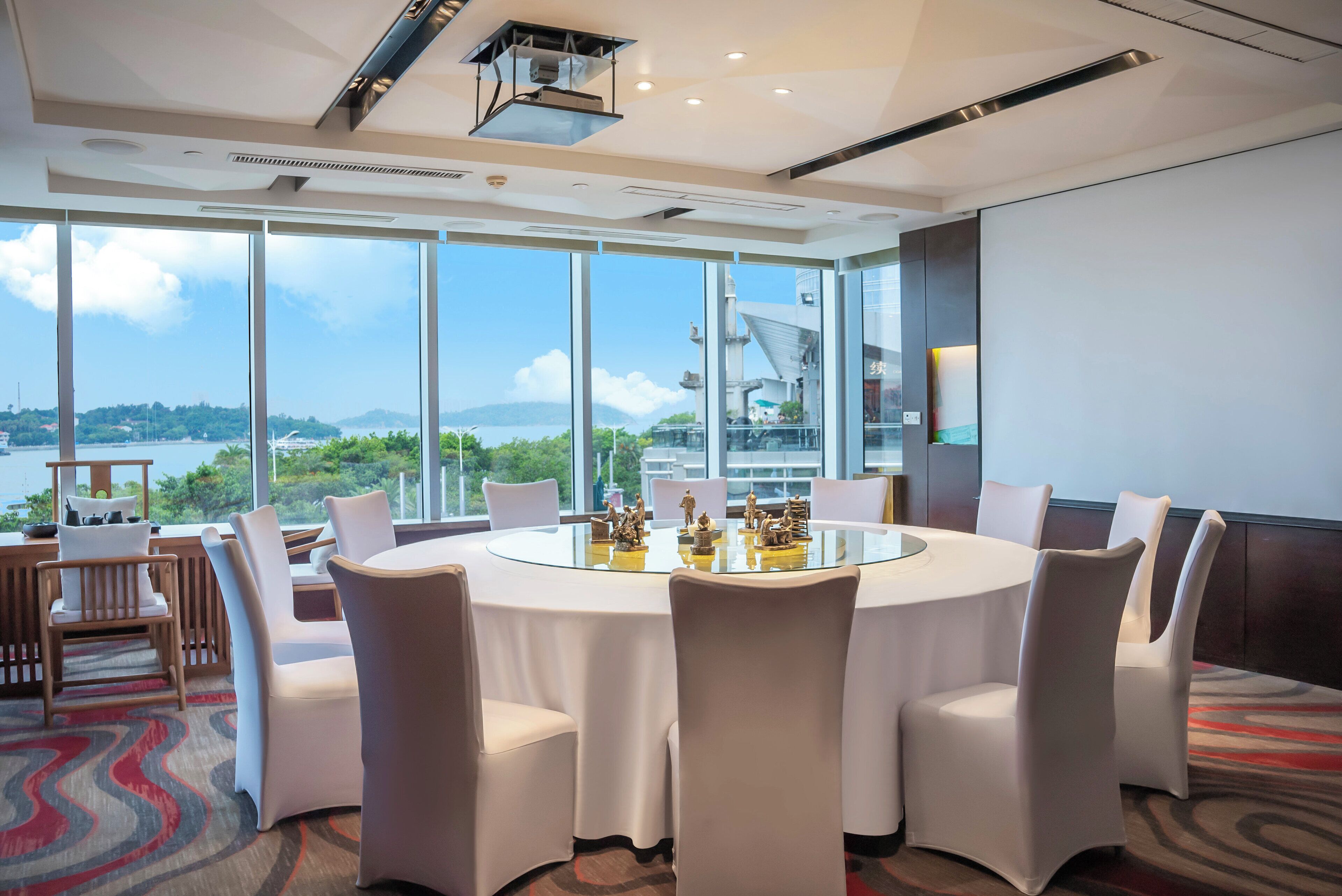 A modern conference room with a glass table surrounded by chairs, large windows with a view of a body of water and greenery, and a projector screen on the wall.