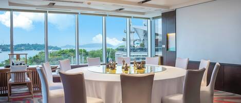 A modern conference room with a glass table surrounded by chairs, large windows with a view of a body of water and greenery, and a projector screen on the wall.