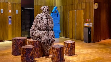 Una escultura de figura humana rodeada de taburetes de madera en un espacio interior con paneles de madera y un ascensor.