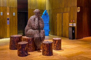 Una escultura de figura humana rodeada de taburetes de madera en un espacio interior con paneles de madera y un ascensor.
