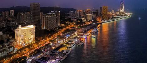 Un paisaje urbano nocturno con un puente destacado, edificios altos y una zona ribereña con barcos.
