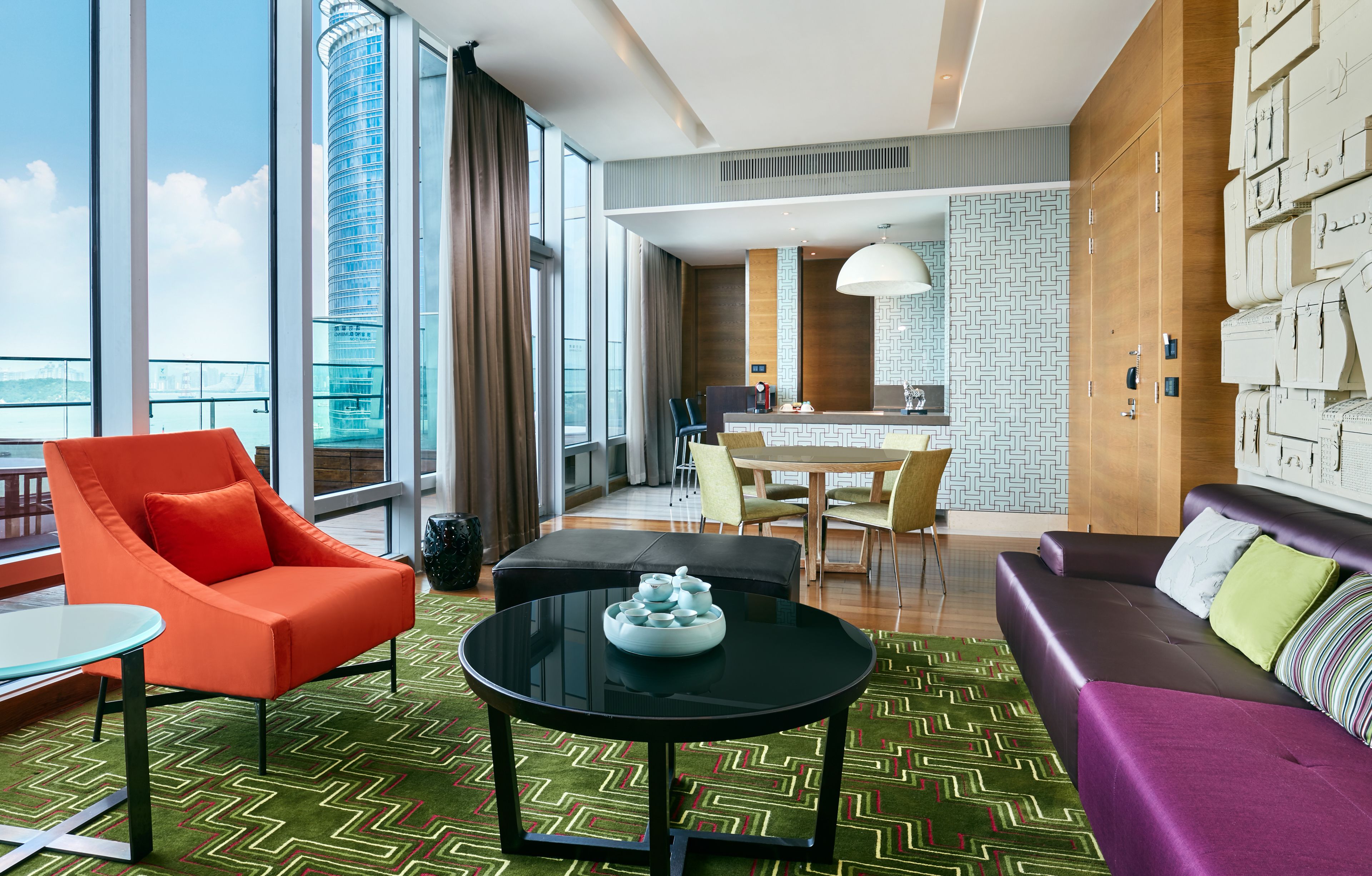 A modern hotel room with a large window, a dining area, and a seating area with a sofa and armchair.