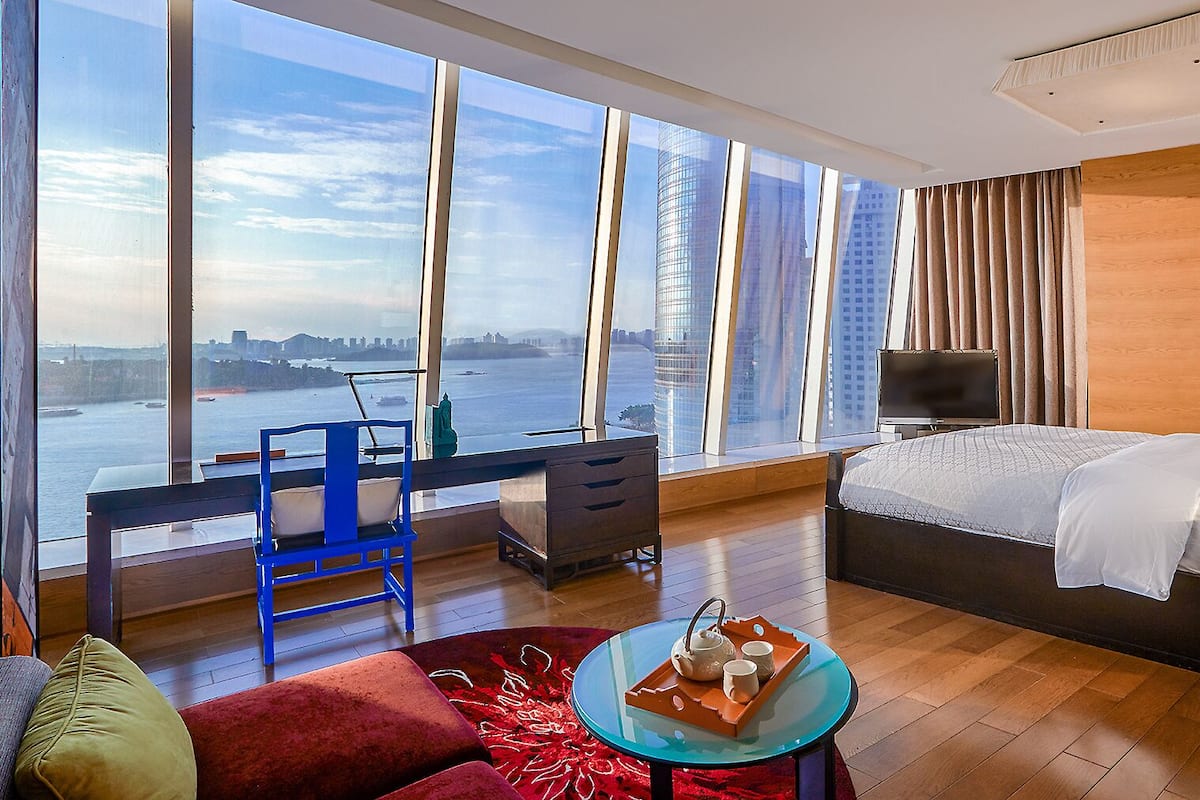 Habitación de hotel con un ventanal grande, cama, escritorio, silla y mesita con bandeja.