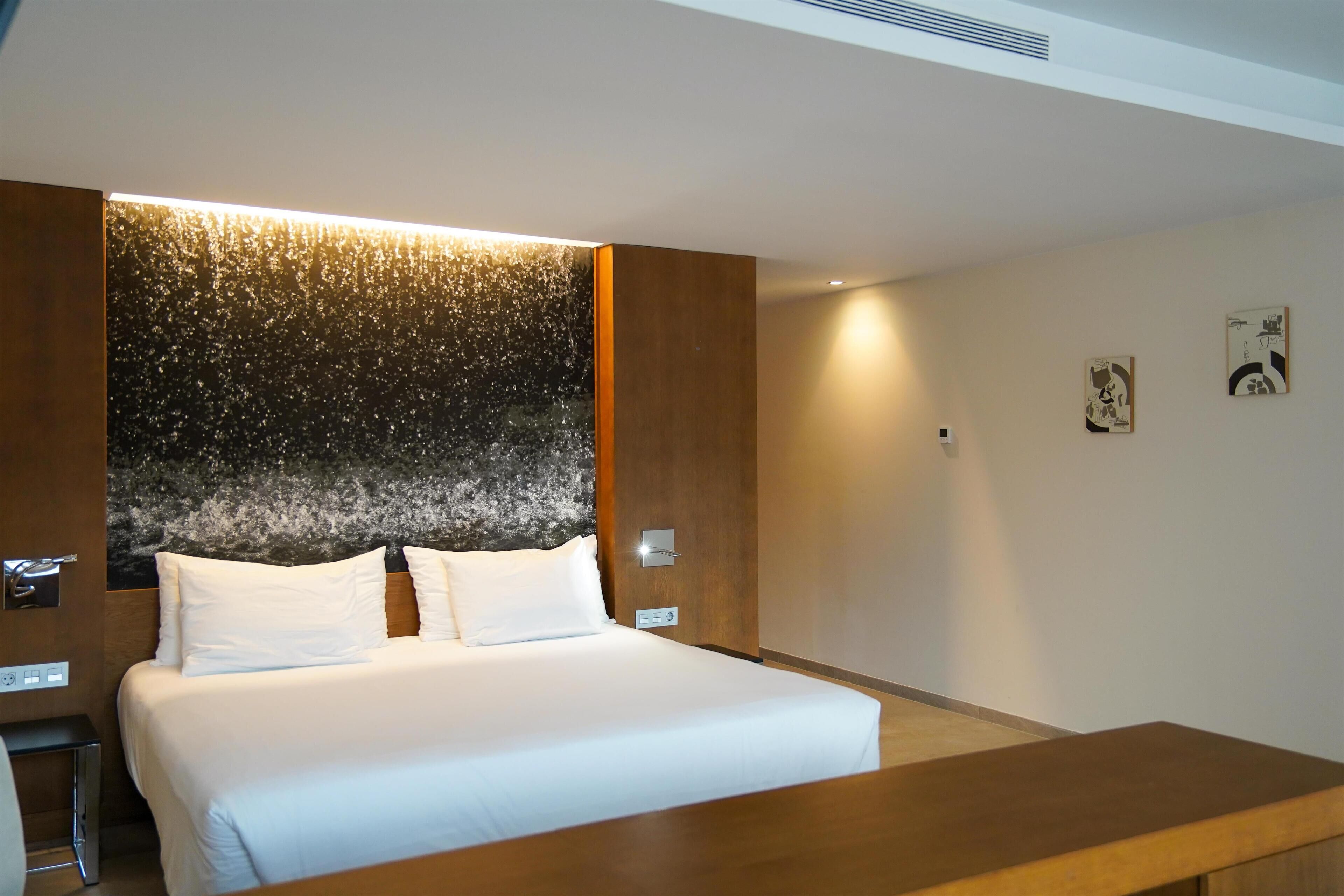 A modern hotel room with a large bed, a wooden headboard, and a wall-mounted light fixture.