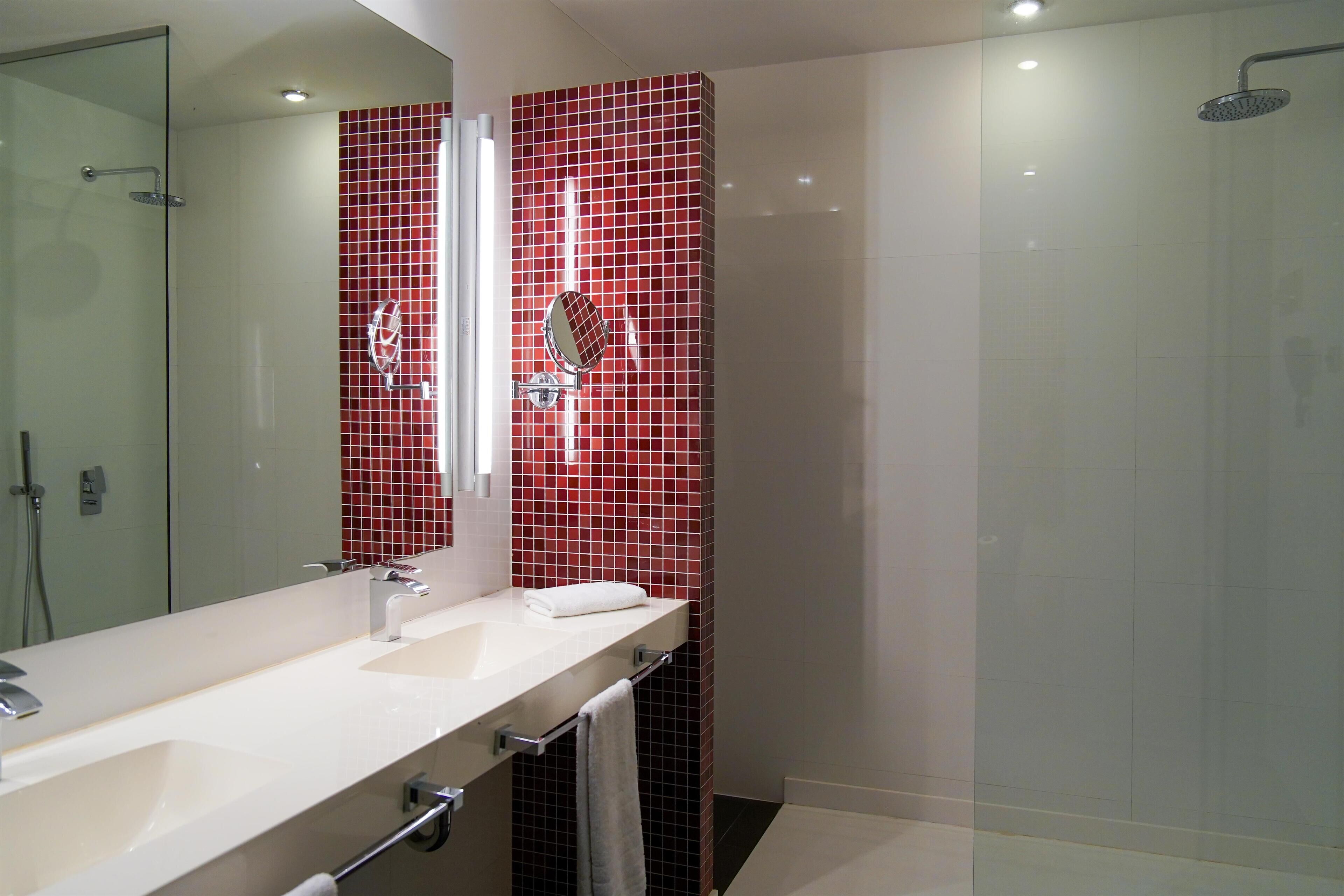 A modern bathroom with a large mirror, two sinks, a glass shower enclosure, and red-tiled walls.