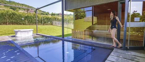 A modern outdoor swimming pool with clear glass walls, a wooden deck, and a view of a green landscape.