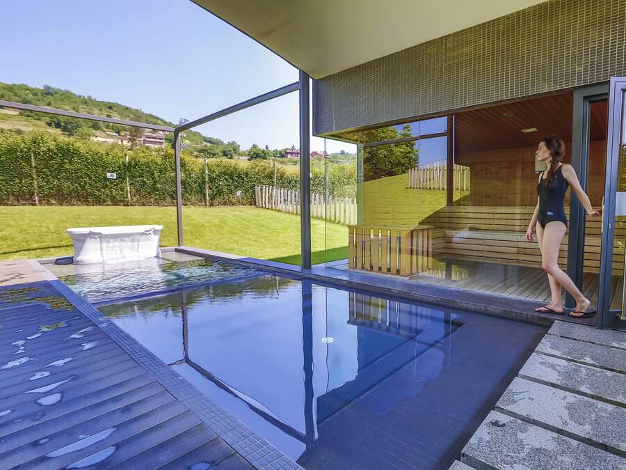 A modern outdoor swimming pool with clear glass walls, a wooden deck, and a view of a green landscape.