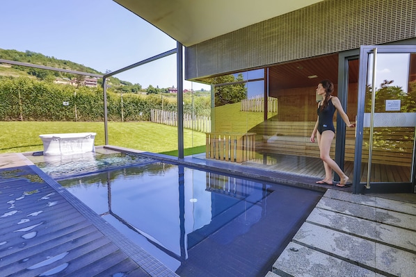 A modern outdoor swimming pool with clear glass walls, a wooden deck, and a view of a green landscape.