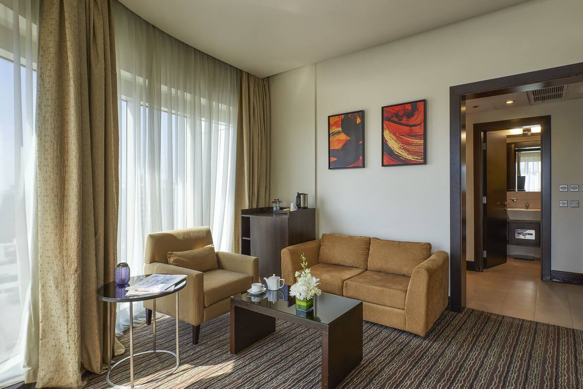 Quarto de hotel com sofá, poltrona, mesa de centro e vista da cidade por grandes janelas.