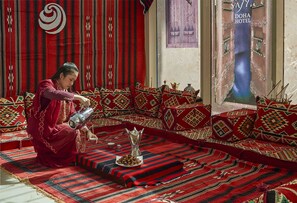 A traditional setting with a person pouring a drink into a cup, a decorative plate with nuts, and a large window with a view of a blue body of water.