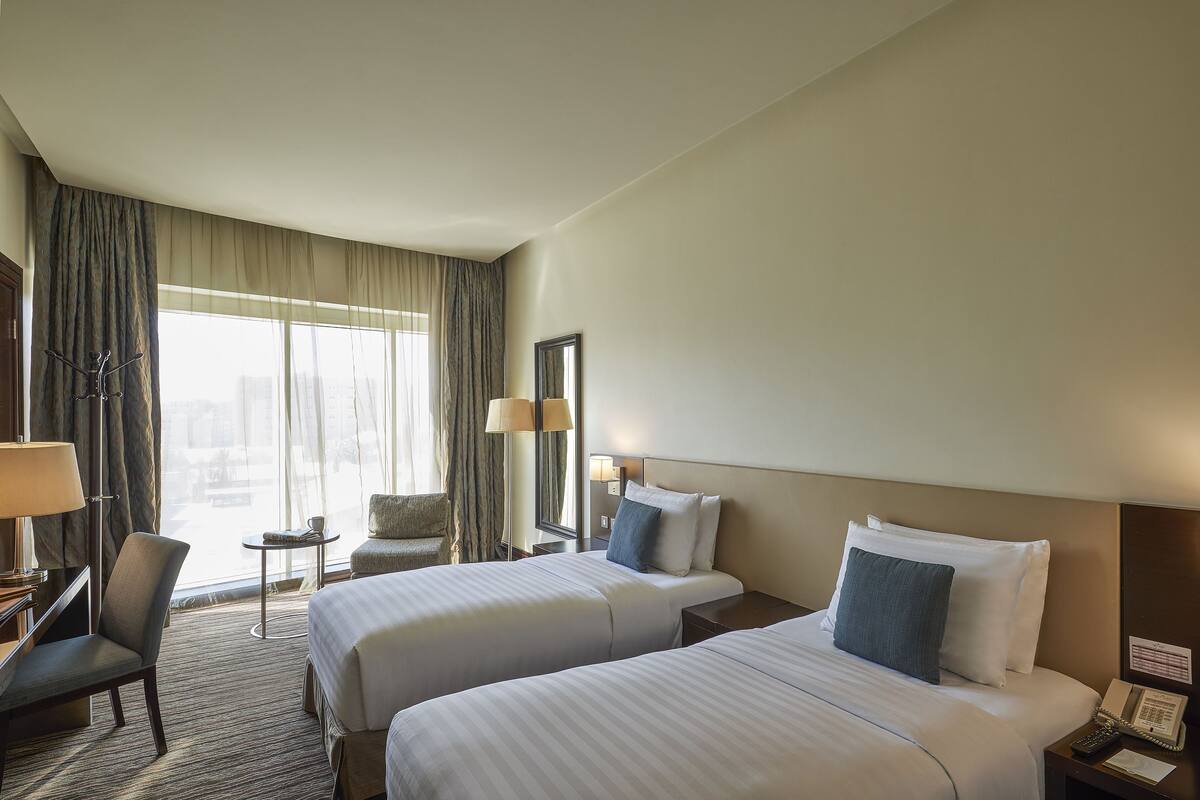 Quarto de hotel com duas camas, uma escrivaninha, uma cadeira, uma mesinha e uma janela com cortinas.