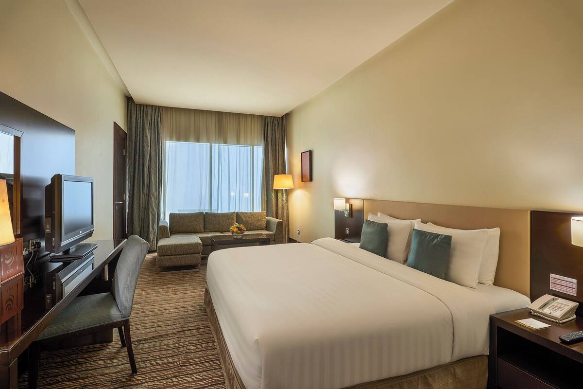 Quarto de hotel com uma cama grande, uma escrivaninha com computador, televisão, sofá e janela com cortinas.