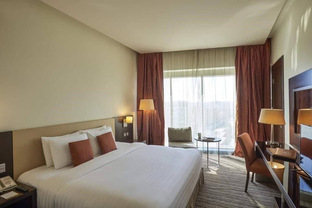 Quarto de hotel com uma cama grande, duas luminárias de cabeceira, uma escrivaninha com cadeira e vista para o exterior pela janela.