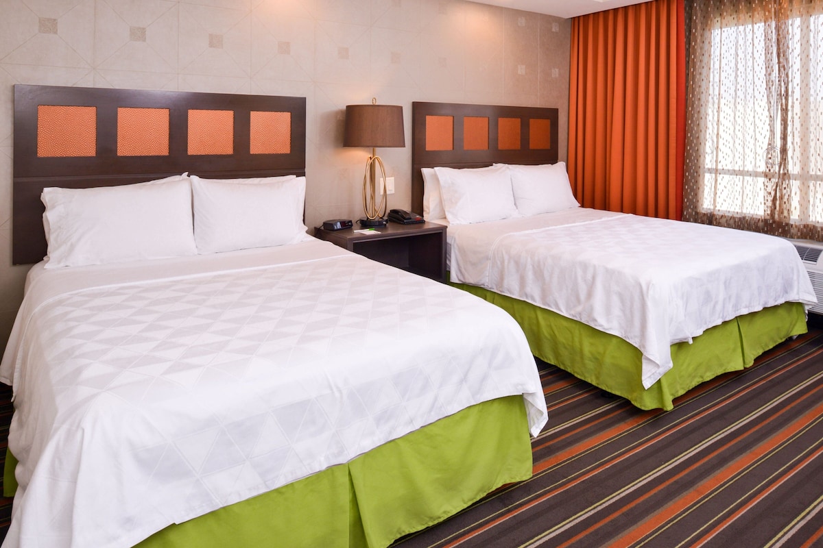 Ein Hotelzimmer mit zwei Betten, einem Nachttisch mit Lampe und einem Fenster mit Vorhängen.