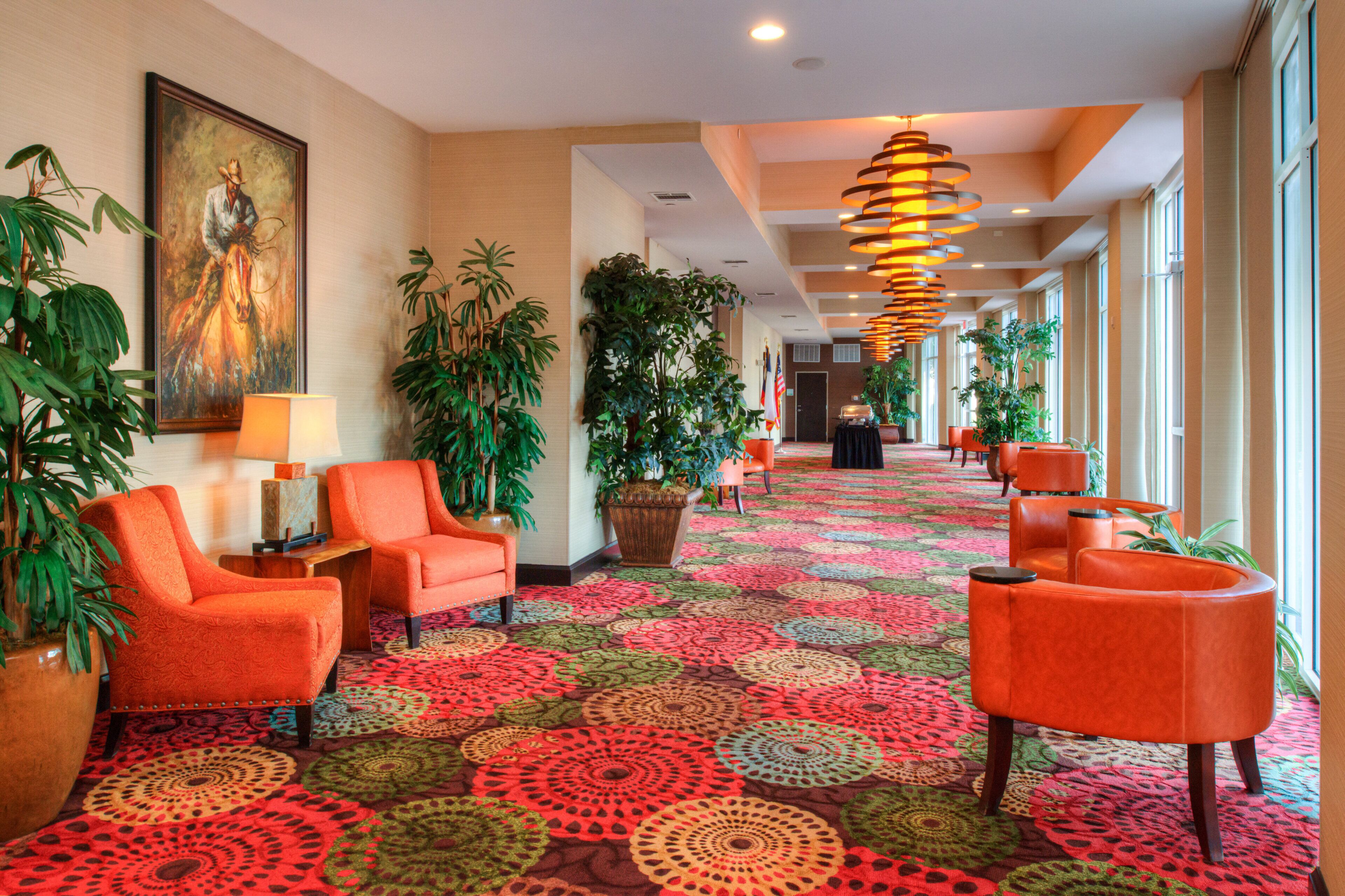 Un hall d’hôtel avec un tapis à motifs, des fauteuils orange, des plantes en pot et un grand tableau accroché au mur.