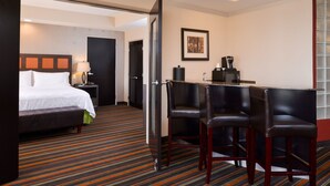 Quarto de hotel com cama, escrivaninha com cadeira, cafeteira e quadro pendurado na parede.
