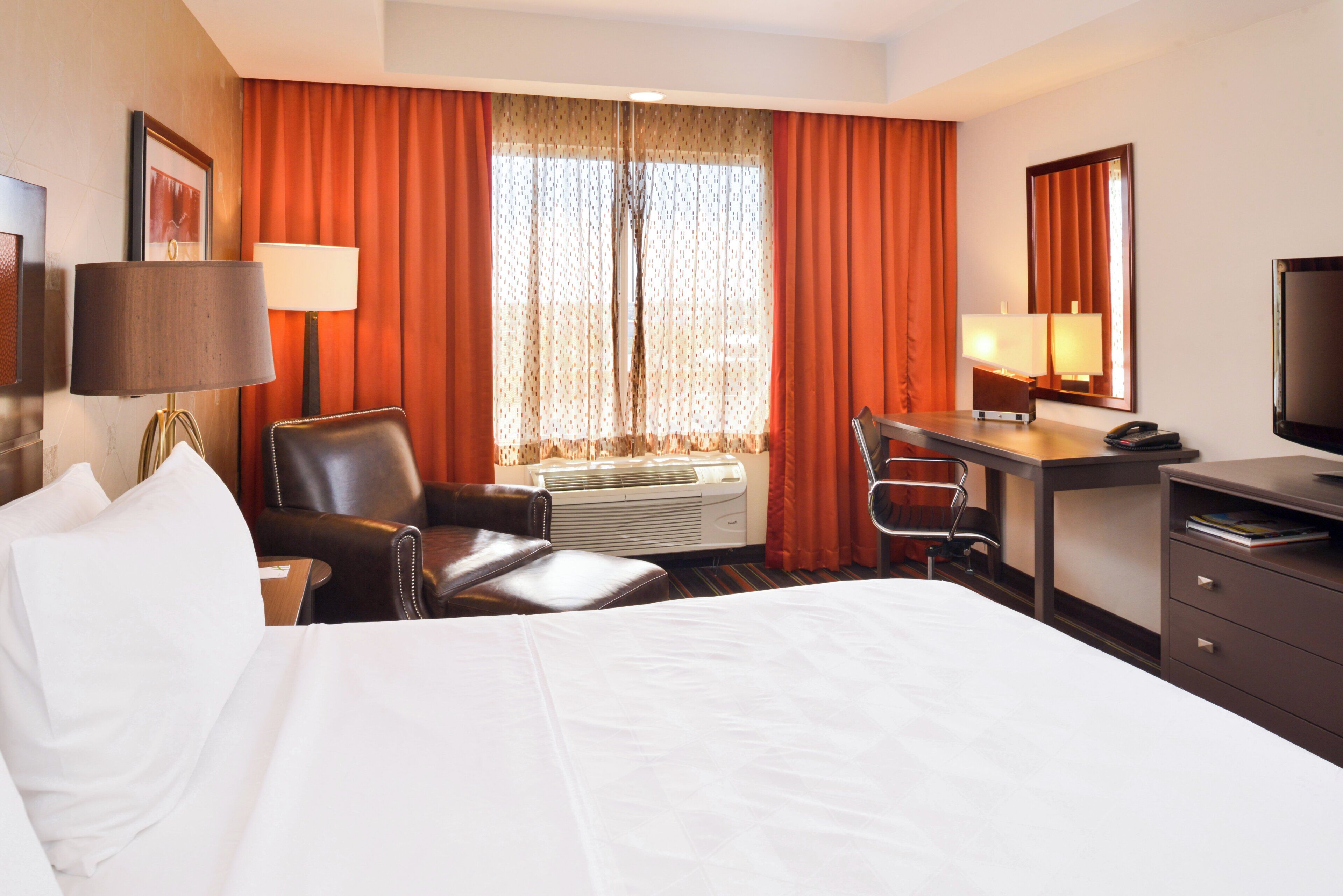 A hotel room with a bed, a desk, a chair, a TV, and an air conditioning unit.