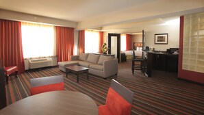 Quarto de hotel com sofá, mesa de jantar, cozinha e uma janela grande com cortinas.