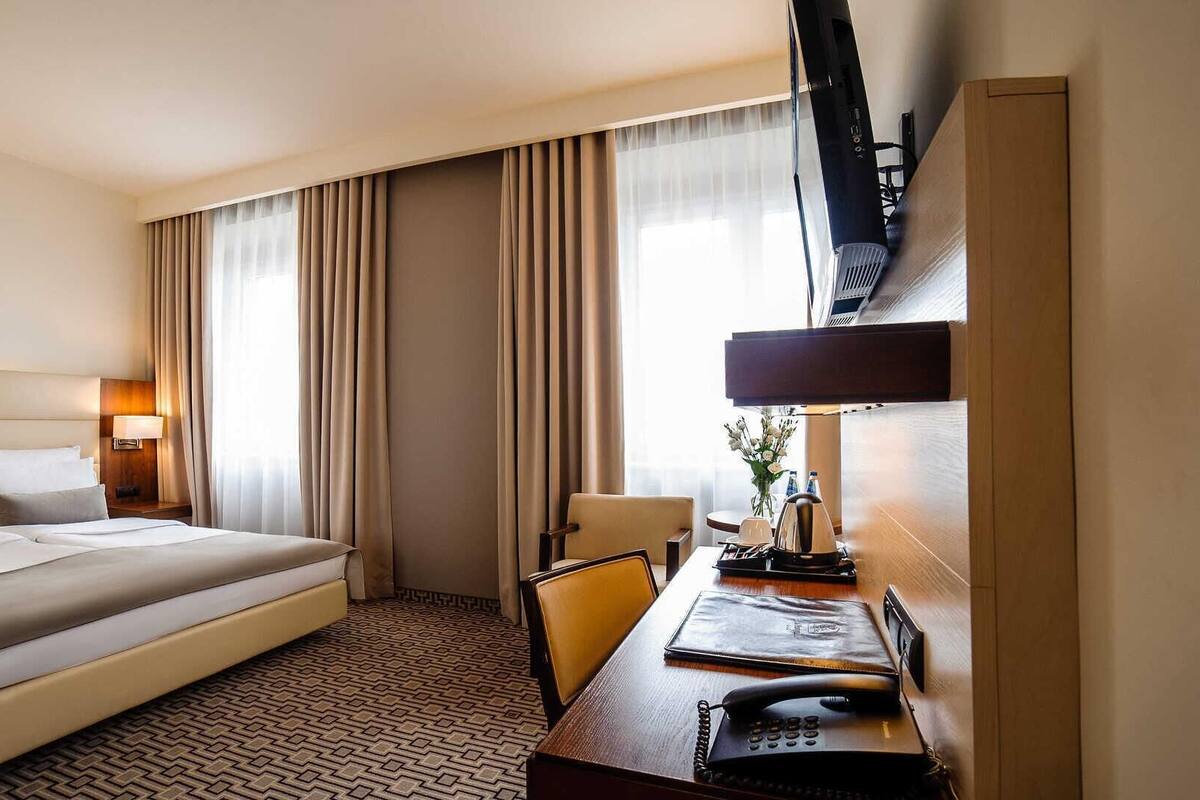 A hotel room with a large bed, a desk with a telephone, a chair, a TV mounted on the wall, and a window with curtains.