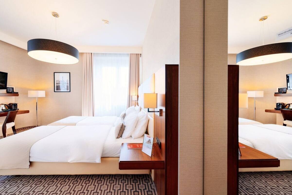 A hotel room with two beds, a desk, a chair, a TV, and a large window.