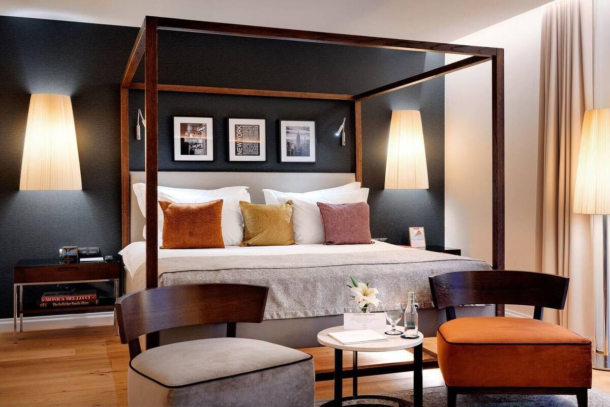 A four-poster bed with a dark headboard, a bedside table with a lamp, and a chair with a cushion.