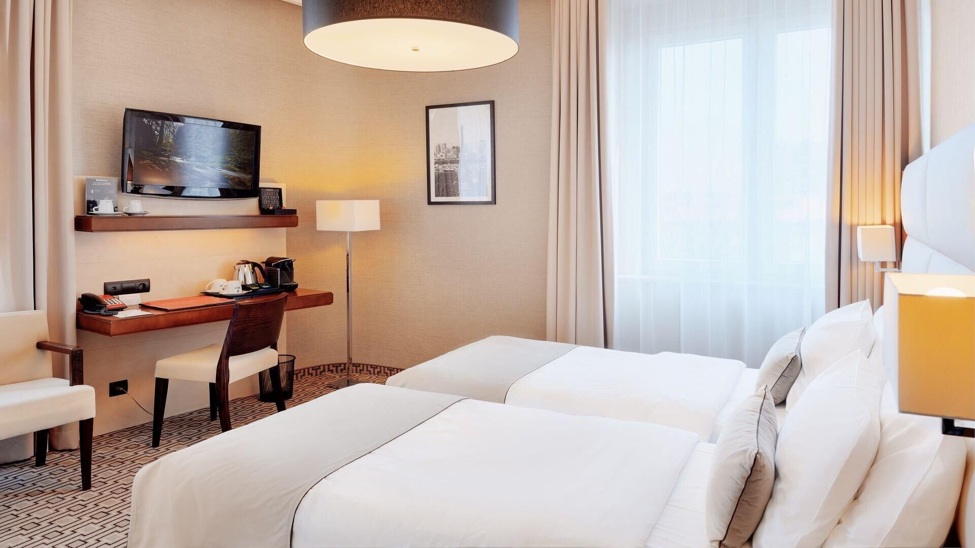 Une chambre d’hôtel avec deux lits, un bureau avec une chaise, une télévision et une fenêtre avec des rideaux légers.