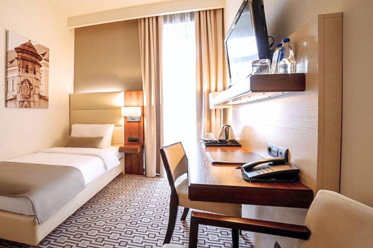 A hotel room with a bed, a desk with a telephone, a chair, a television, and a window with curtains.