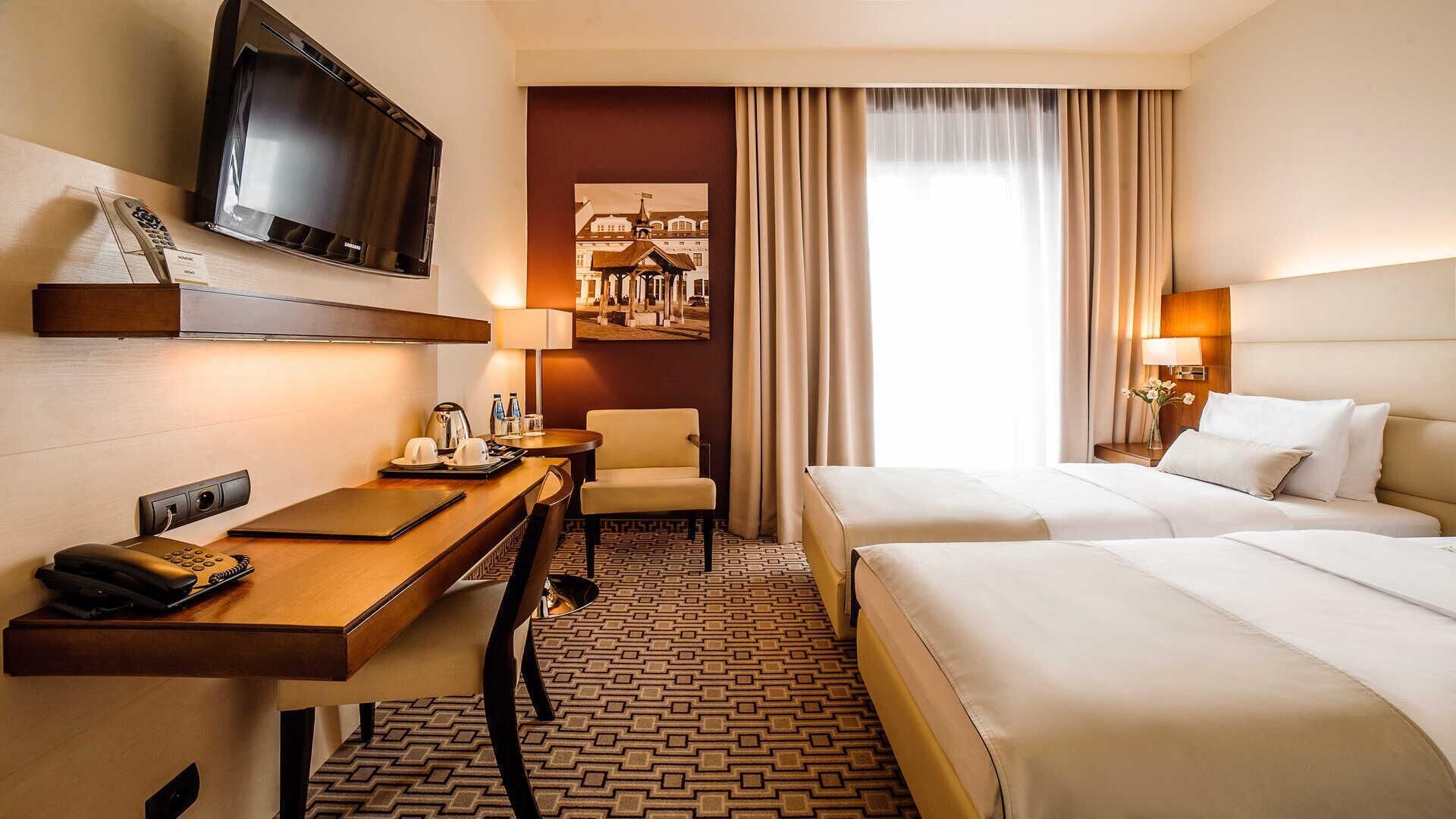 Una habitación de hotel con una cama, un escritorio con teléfono, una silla, un televisor montado en la pared y un cuadro de un edificio.