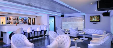 A modern lounge area with white leather sofas and chairs, a bar with liquor bottles, and a television mounted on the wall.
