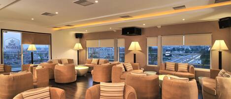 A spacious lounge area with multiple sofas and armchairs, large windows offering city views, and ambient lighting.