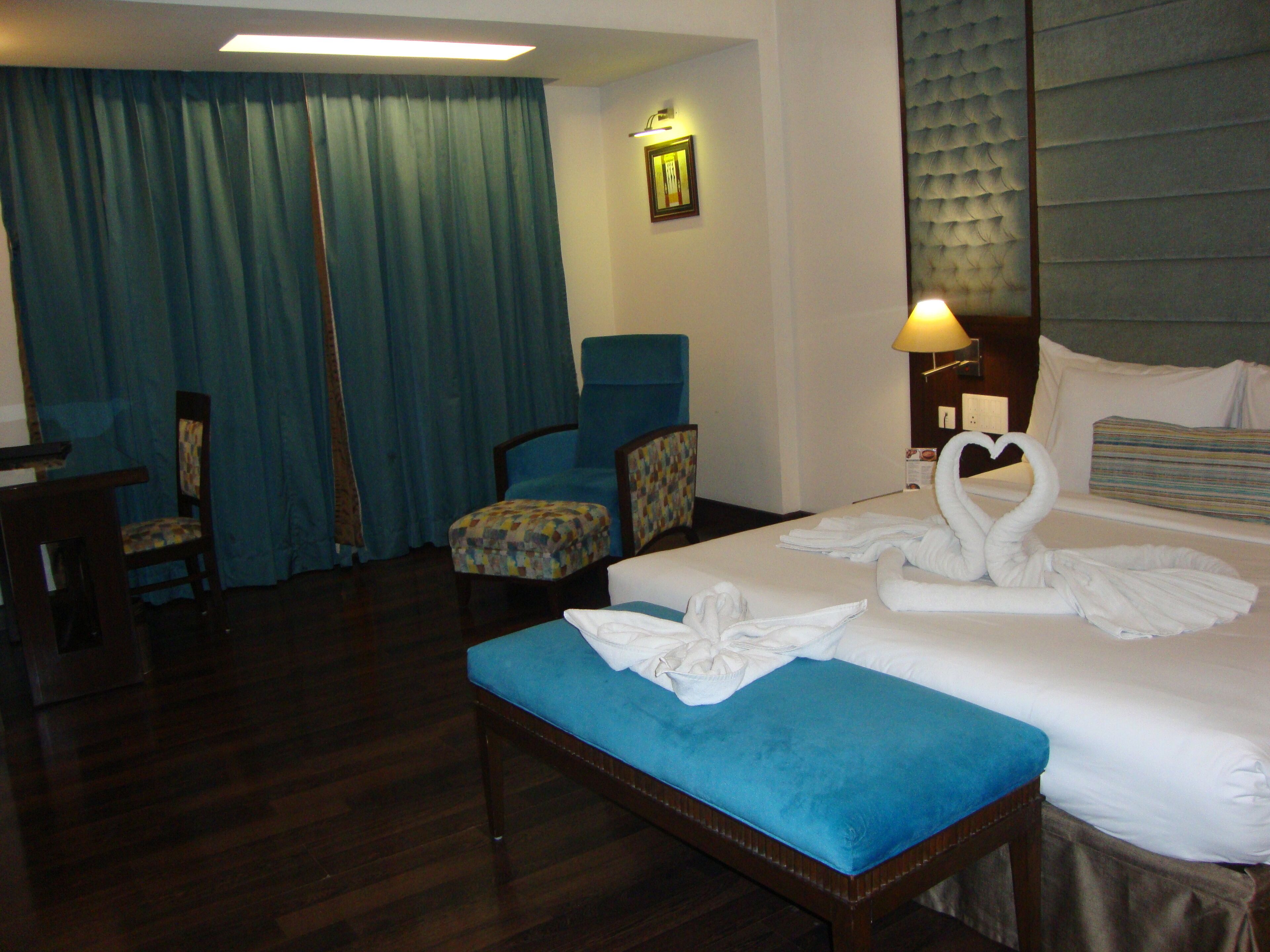 Una habitación de hotel con una cama, un banco tapizado en azul, una silla, un escritorio y un ventanal con cortinas azules.