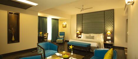 A hotel room with a bed, two blue armchairs, a coffee table with a vase and fruit, and a nightstand with a lamp.